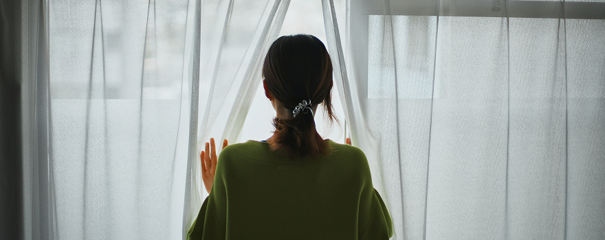窓辺のカーテンを開けて外を見る女性の後ろ姿の写真